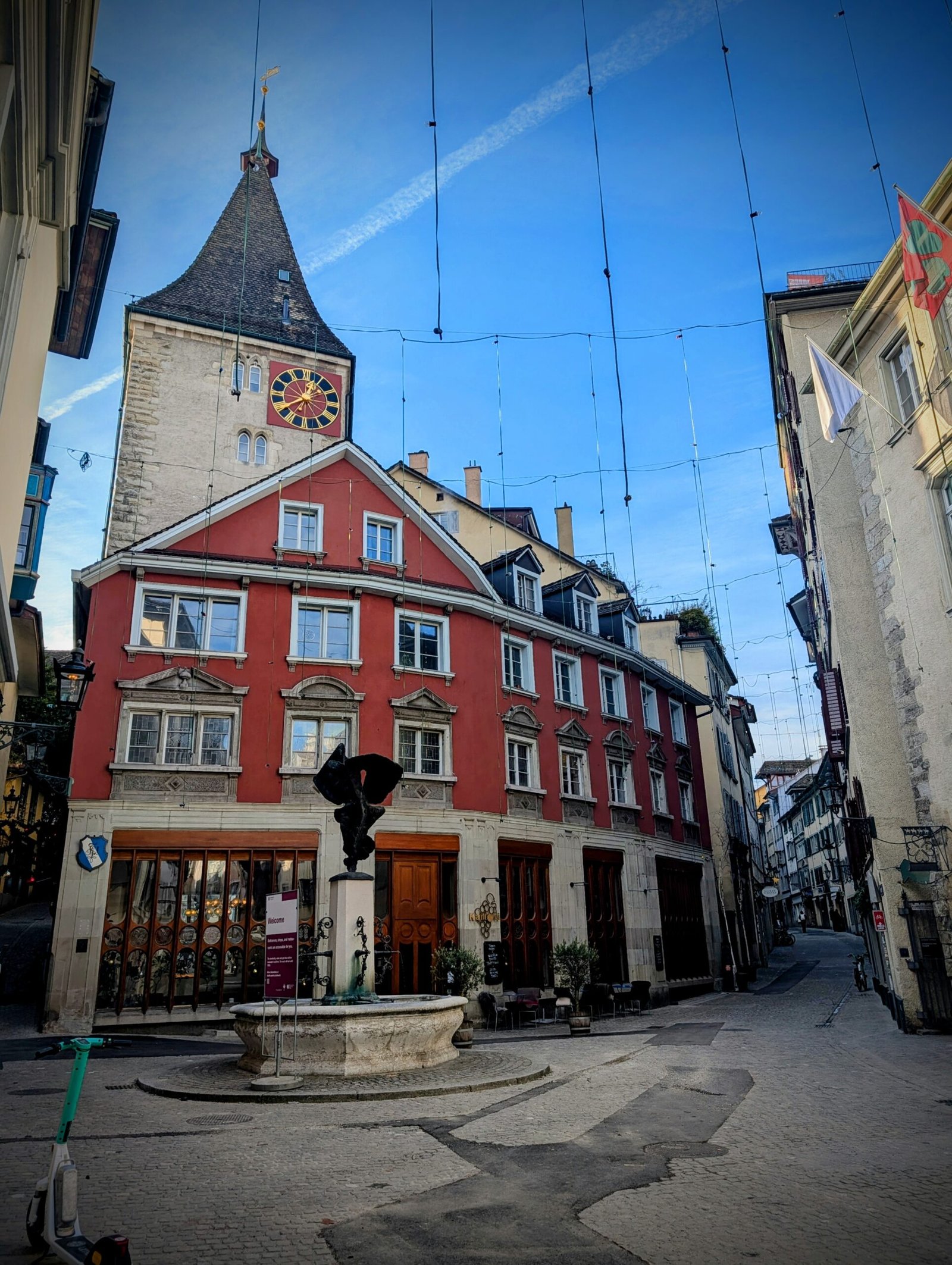Grimmenturm clock tower and historic red building in Zurich Old Town, viewed along a quiet cobblestone street.