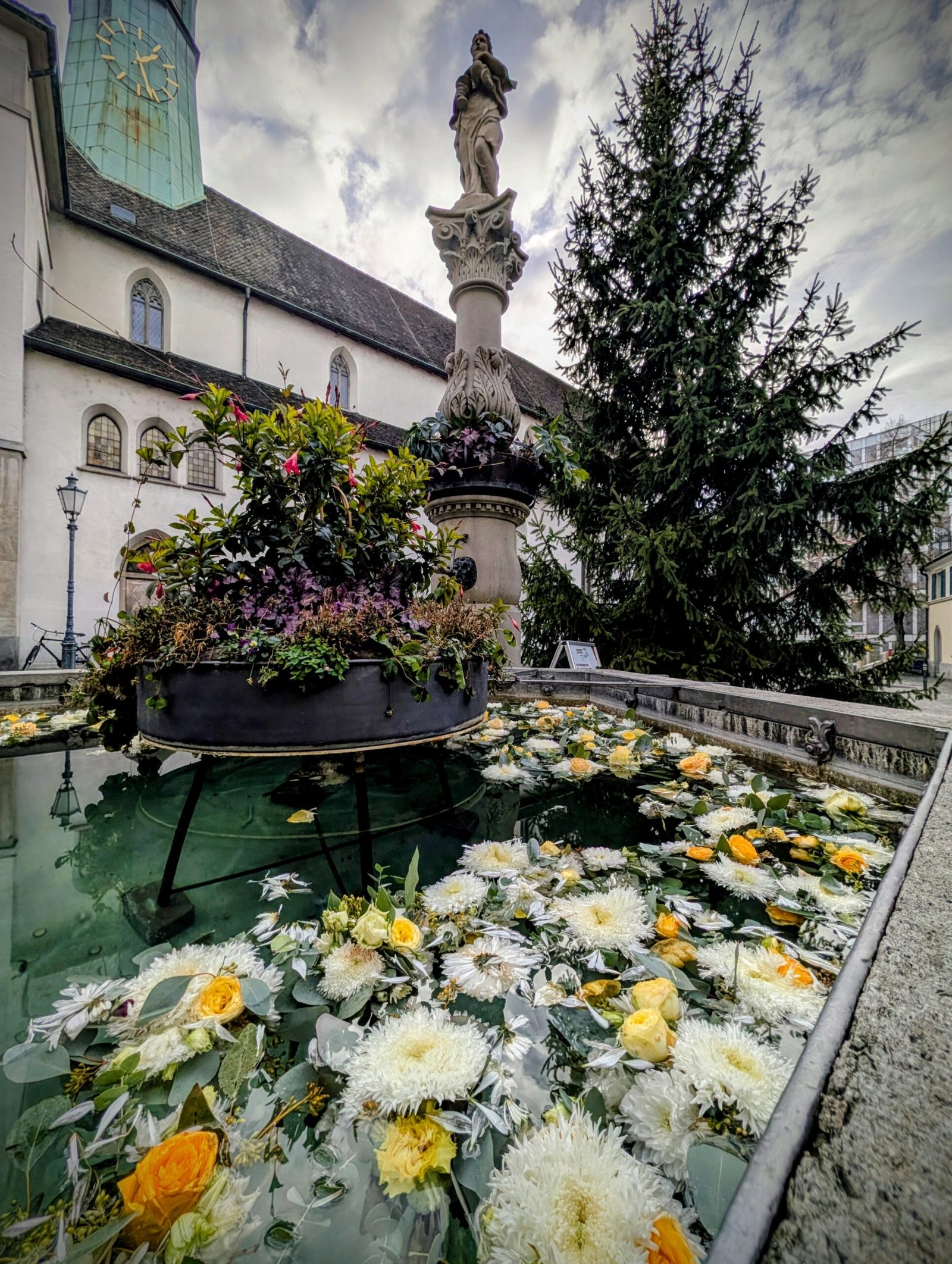 Flower-filled fountain along Augustinergasse in Zurich Old Town, with a stone statue rising above the water and historic buildings in the background.