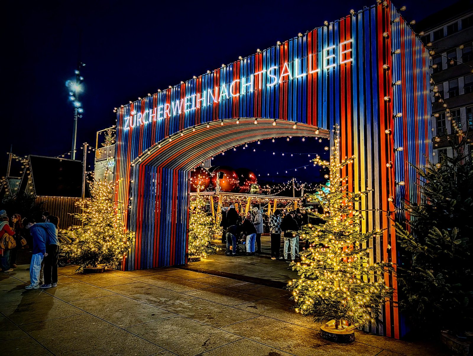 Illuminated entrance to the Zurich Christmas Market at night, with festive lights, decorated trees, and visitors walking through.