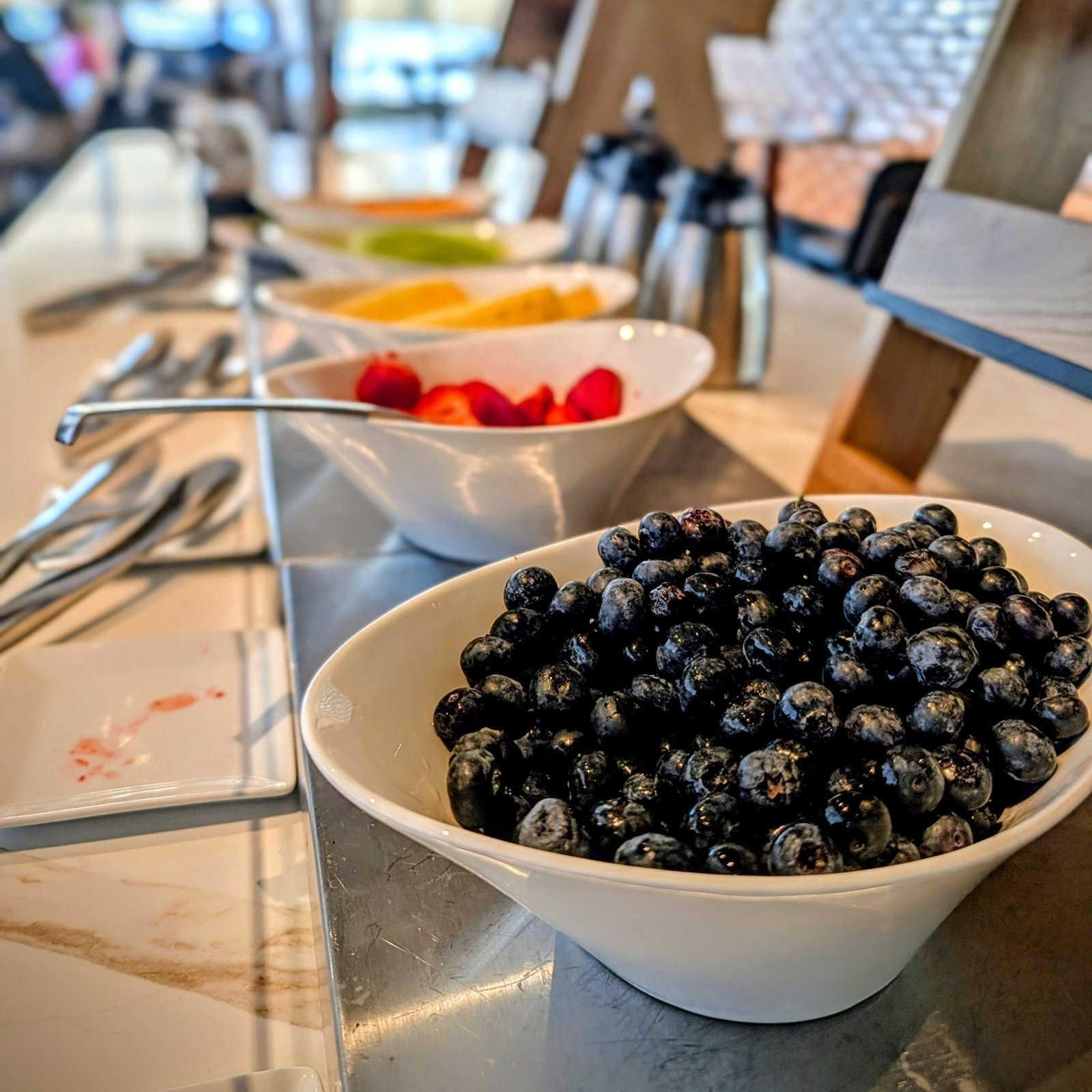 Bowls of fresh blueberries, strawberries, and other fruit arranged on a hotel breakfast buffet counter with serving utensils nearby.